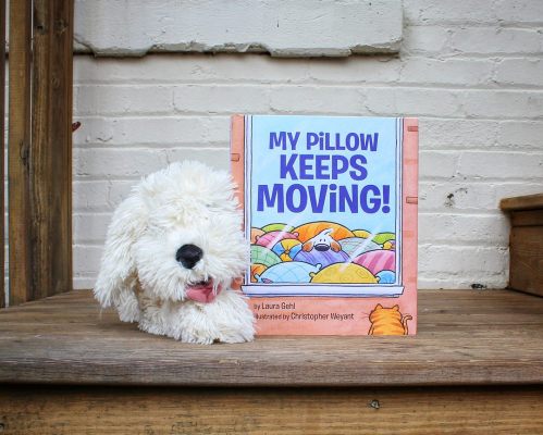 A fluffy dog toy sits beside the children's book "My Pillow Keeps Moving!" on a wooden surface.