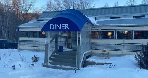 A diner entrance with a blue awning, surrounded by snow and trees, showcasing a cozy atmosphere.