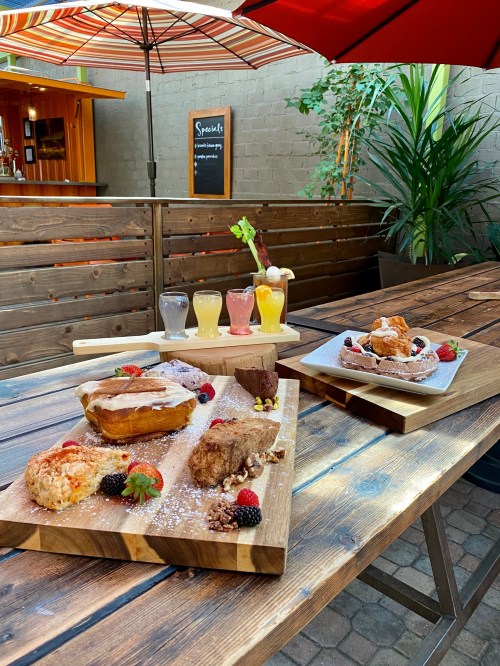 A wooden table outdoors displays a variety of colorful drinks and delicious desserts, surrounded by greenery and umbrellas.