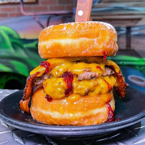 A towering burger made with two glazed donuts, a beef patty, melted cheese, bacon, and a drizzle of sauce.