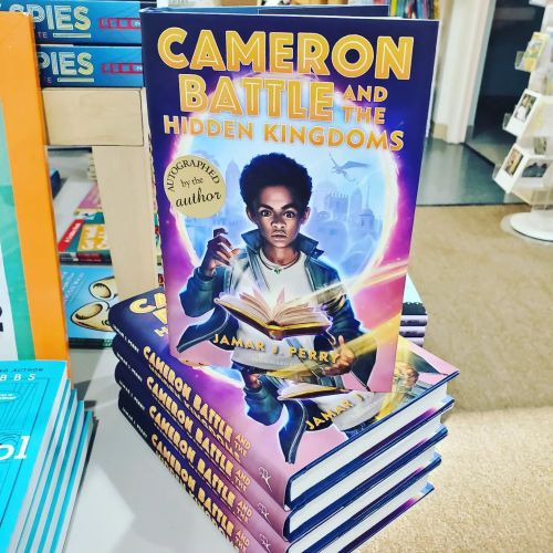 A stack of books titled "Cameron Battle and the Hidden Kingdoms" by Jamari Perry, featuring a young boy holding a book.