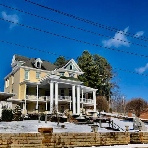 A large yellow house with white columns, surrounded by snow, under a clear blue sky and tall trees in the background.