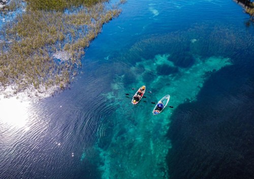 Two kayakers paddle in clear blue water surrounded by lush greenery and sunlight reflecting on the surface.