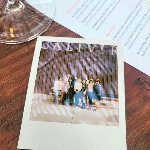 A group of people posing together in front of a sign, with a menu and a glass in the foreground.