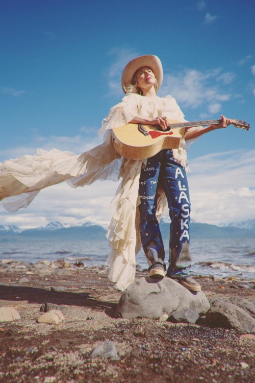 A musician in a flowing outfit stands on a rock by the beach, playing guitar against a scenic backdrop.