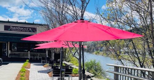 Bright pink umbrellas shade outdoor seating by a river, with a restaurant in the background on a sunny day.