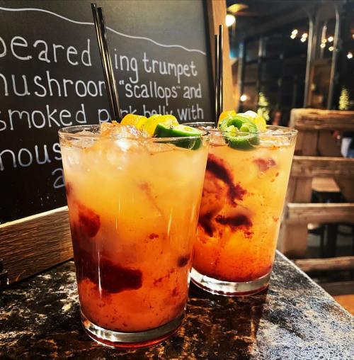 Two colorful cocktails with ice, garnished with lime and orange slices, sit on a table next to a chalkboard menu.