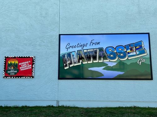 Mural on a wall reading "Greetings From Hiawassee, GA" alongside a sign featuring the town's name and founding year.