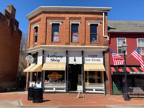 A brick storefront with striped awnings, featuring "The Lollipop Shop" sign, surrounded by American flags and a clear blue sky.