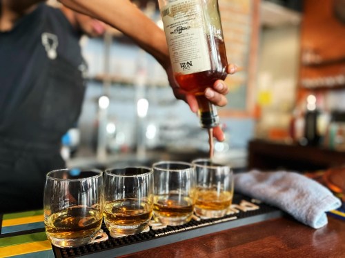 A bartender pours whiskey into four glasses lined up on a bar counter.
