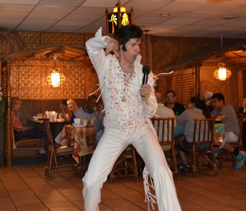 A performer in a white jumpsuit sings passionately on stage, with an audience enjoying the show in a lively restaurant.