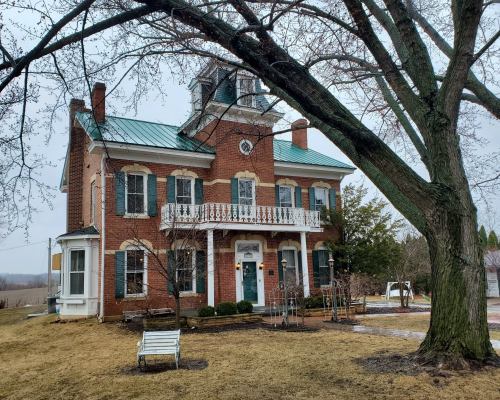 A historic brick house with a green metal roof, large windows, and a porch, surrounded by bare trees and a grassy yard.