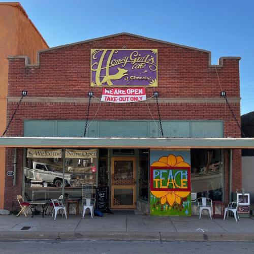 A brick storefront with a colorful sign reading "Honey Girl's Cafe & Chocolate," featuring outdoor seating and a peace mural.