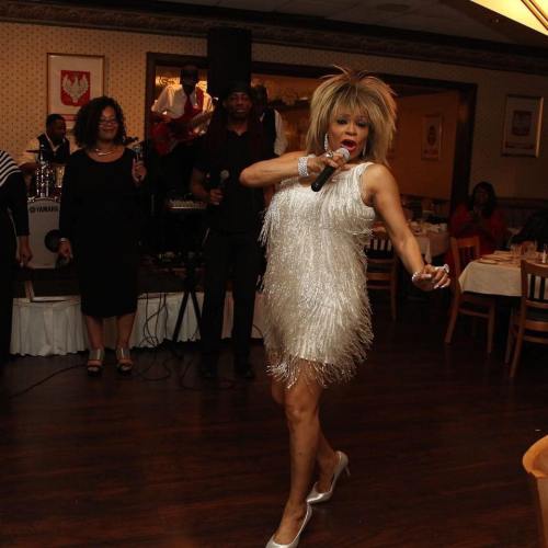 A performer in a sparkly dress sings on stage, with musicians and an audience in a lively restaurant setting.