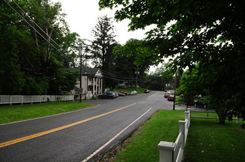 A quiet street lined with trees, featuring parked cars and a building on the left, under a cloudy sky.