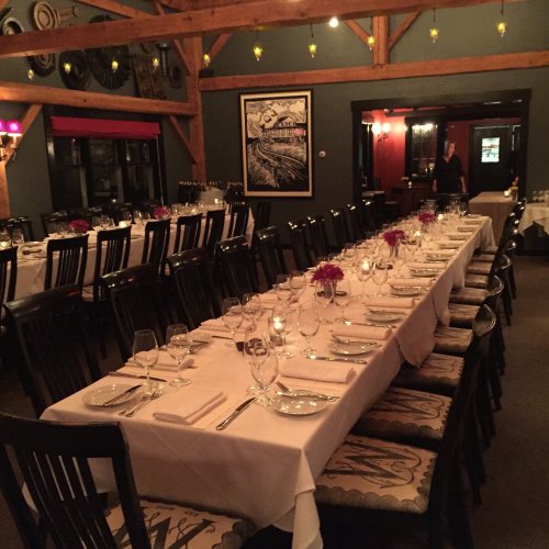 A warmly lit dining room with long tables set for a formal dinner, featuring candles and floral centerpieces.