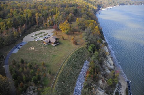Aerial view of a house on a cliff overlooking a river, surrounded by colorful autumn trees and a winding driveway.