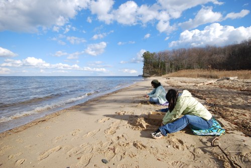 Two people sit on a sandy beach, focused on the water, with trees and clouds in the background.