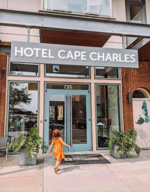A child in an orange dress walks towards the entrance of Hotel Cape Charles, featuring a welcoming sign and plants.