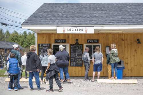 A group of people waiting in line at a dairy bar with a wooden exterior and a menu board.