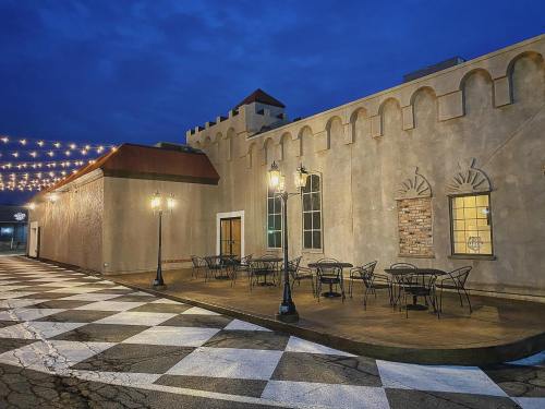 A charming outdoor seating area with black chairs and tables beside a textured building, illuminated by string lights at dusk.