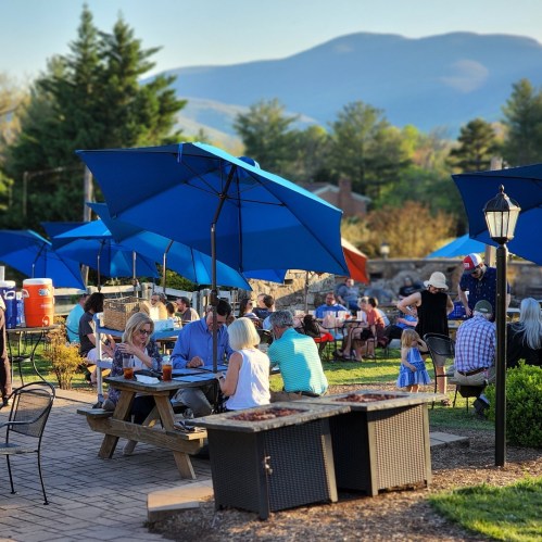 Outdoor dining scene with blue umbrellas, people enjoying meals, and mountains in the background on a sunny day.