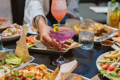 A table filled with various dishes and drinks, featuring a hand holding a purple cocktail with a lemon garnish.