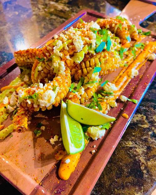 A wooden platter with grilled corn topped with cheese, herbs, and lime wedges, drizzled with sauce.