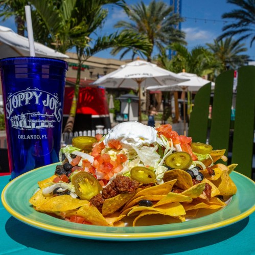 A colorful plate of nachos topped with cheese, jalapeños, lettuce, tomatoes, and sour cream, with a drink in the background.