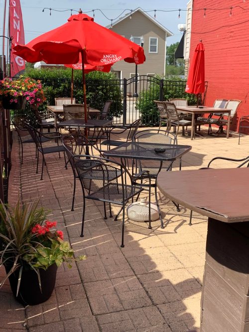 Outdoor patio with tables and chairs, red umbrellas, and potted plants, surrounded by greenery and a red wall.