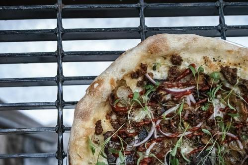 A close-up of a pizza topped with sausage, pickles, onions, and herbs on a grill grate.
