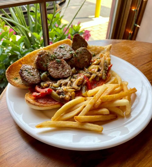 A plate of sausage sandwich with sautéed vegetables and fries, set against a backdrop of greenery.
