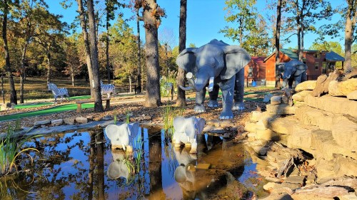 A serene scene featuring two elephant sculptures by a pond, surrounded by trees and zebra decorations in the background.