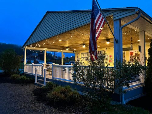 A cozy storefront illuminated at dusk, featuring string lights and an American flag, surrounded by greenery.