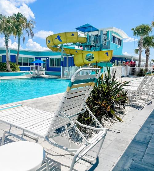 A sunny pool area with white lounge chairs, a water slide, and a colorful building in the background. Palm trees are visible.