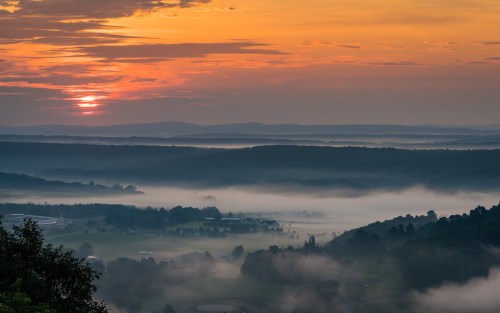 A serene sunrise over misty valleys, with layers of hills and soft clouds illuminated by warm orange and yellow hues.