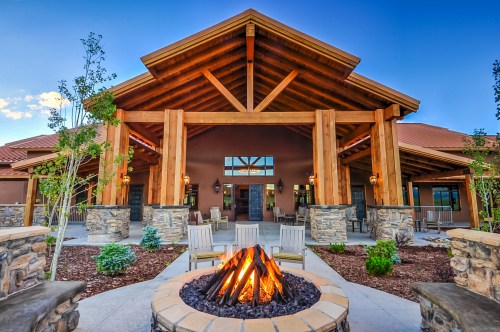 A cozy outdoor space featuring a stone fire pit, wooden beams, and lush greenery in front of a welcoming lodge.