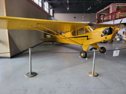A vintage yellow airplane displayed indoors, with a propeller and a chain barrier around it.