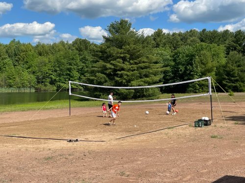 A group of children playing volleyball on a sandy court near a lake, surrounded by trees and a blue sky.