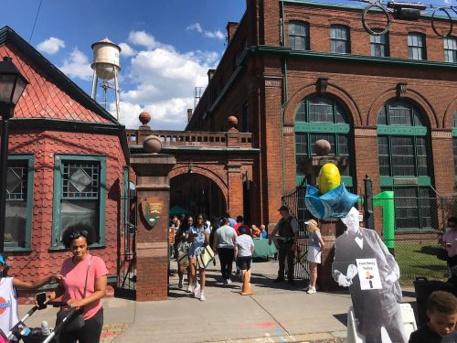 A bustling outdoor event with people entering a brick building, featuring a cutout figure holding a sign.