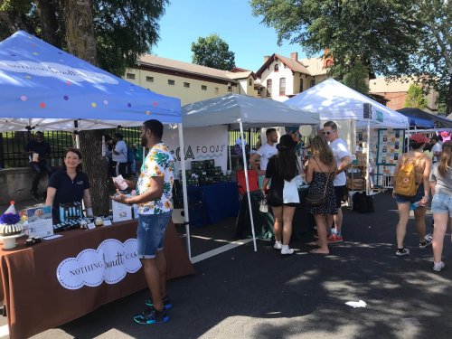 A bustling outdoor market with vendor tents, people browsing, and a variety of products on display.