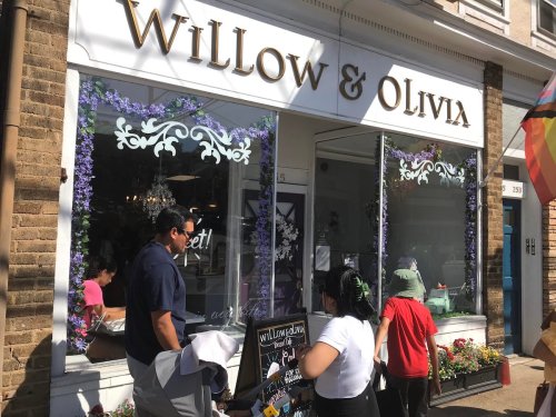 A storefront with "Willow & Olivia" sign, featuring floral decorations and people outside, enjoying the sunny day.