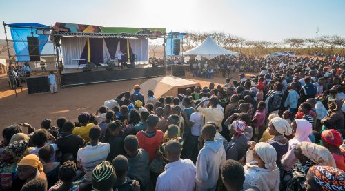 A large crowd gathers at an outdoor stage for a performance, with tents and a clear sky in the background.