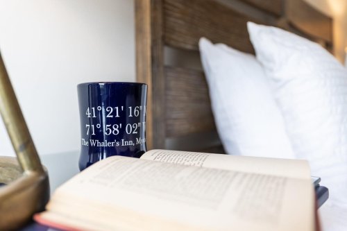 A navy mug with coordinates sits next to an open book on a bedside table, with pillows in the background.