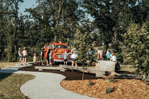 A group of people gathers around a vintage orange truck in a park, surrounded by trees and a winding path.