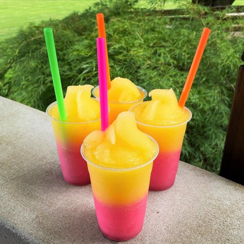 Four colorful slushie cups with yellow and pink layers, topped with swirls, sit on a table with green foliage in the background.