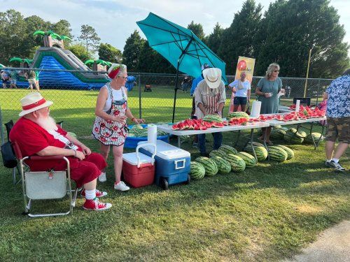 A festive scene with Santa sitting beside a table of watermelons, while people enjoy a summer event in a park.