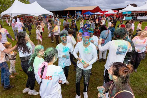 A vibrant outdoor festival scene with people covered in colorful powder, dancing and celebrating under cloudy skies.