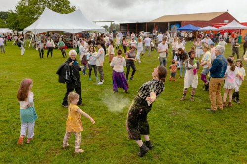 A lively outdoor festival scene with people dancing and celebrating, covered in colorful powder, under a cloudy sky.
