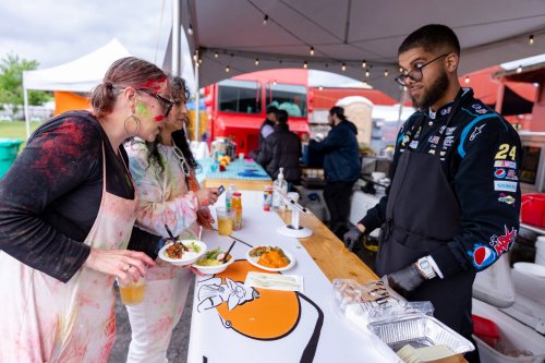 Three people enjoy food at a food stall, while a vendor in a racing suit serves them under a tent.